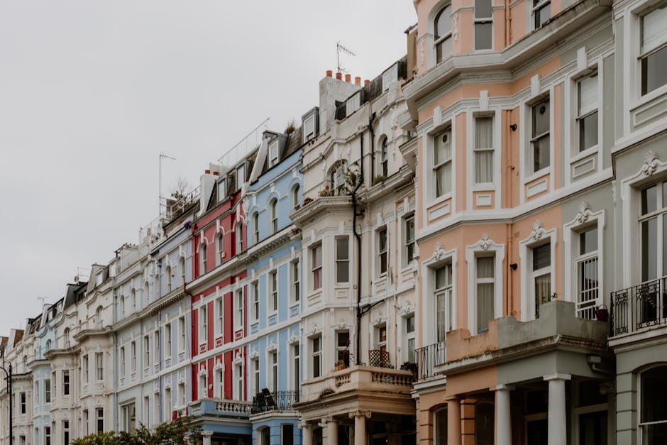 A row of Victorian-style terraced houses in Notting Hill, London, featuring ornate bay windows and decorative moldings. The buildings are painted in pastel shades including light pink, cream, white, red, and blue, with black wrought-iron balconies. The overcast sky casts diffuse lighting over the scene, highlighting the subtle textures of the facades and the variety of architectural details. The street below is not visible, but the scene suggests a typical residential area with a cohesive yet colorful streetscape. The image emphasizes the architectural charm and uniformity of the neighborhood, consistent with the style seen in Notting Hill, with no visible gardens or outdoor maintenance activities. This setting contrasts with typical gardening scenes but adds context to the local environment where outdoor care could be part of overall property upkeep, which a company like Gardeners Notting Hill might service.