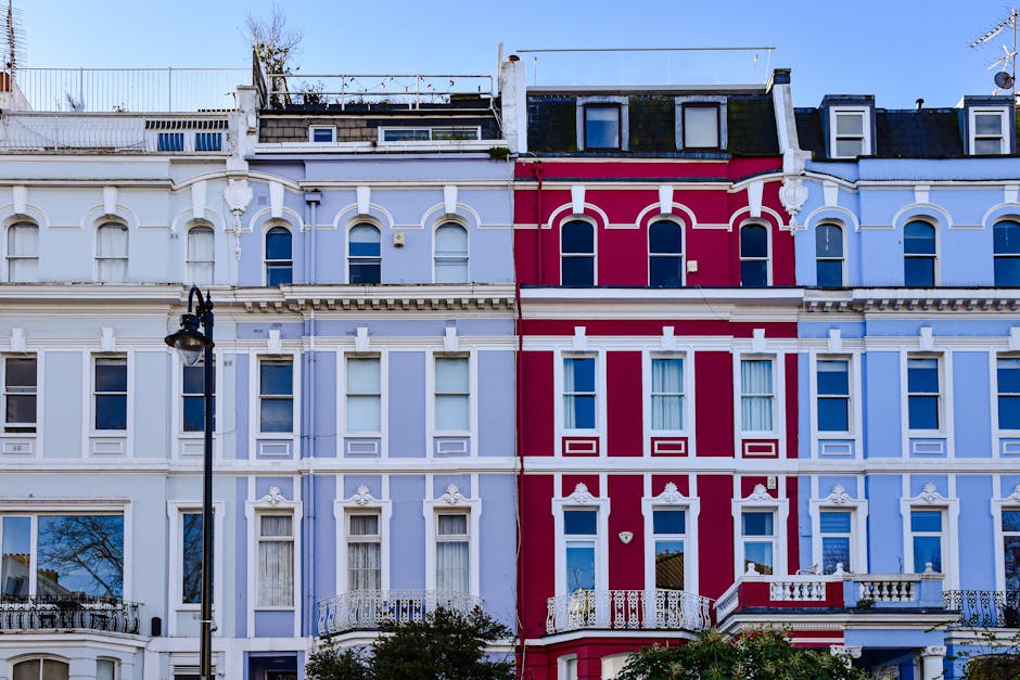 A row of Victorian terraced buildings with ornate architectural details and decorative moldings, painted in shades of white, light blue, and bright pink, with multiple windows featuring white trim. The buildings have small balconies and roof terraces with fencing, and a traditional black street lamp is visible in the foreground. The sky above is clear and blue, providing natural lighting that highlights the textures of the painted facades and the overall urban landscape. This image showcases the characteristic exterior features of Notting Hill architecture, with well-maintained facades that reflect the vibrant and diverse aesthetic typical of the area, fitting for gardening and outdoor maintenance themes related to property upkeep and house exterior care.