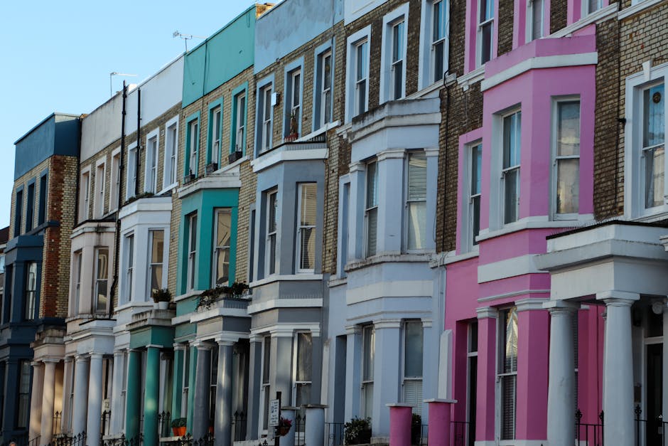 A row of Victorian-style terraced houses in Notting Hill, London, featuring ornate bay windows and decorative moldings. The buildings are painted in pastel shades including light pink, cream, white, red, and blue, with black wrought-iron balconies. The overcast sky casts diffuse lighting over the scene, highlighting the subtle textures of the facades and the variety of architectural details. The street below is not visible, but the scene suggests a typical residential area with a cohesive yet colorful streetscape. The image emphasizes the architectural charm and uniformity of the neighborhood, consistent with the style seen in Notting Hill, with no visible gardens or outdoor maintenance activities. This setting contrasts with typical gardening scenes but adds context to the local environment where outdoor care could be part of overall property upkeep, which a company like Gardeners Notting Hill might service.