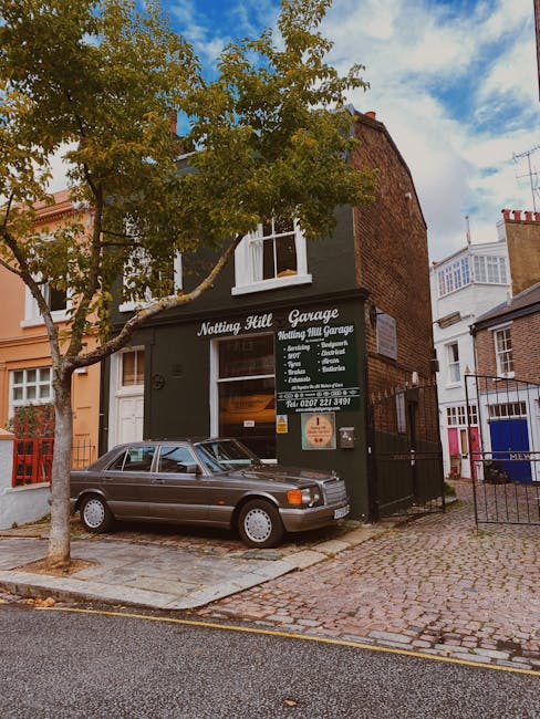 A small urban garden located in front of a black-painted garage building in Notting Hill, W11. The garden features a mature deciduous tree with a textured trunk and green foliage, providing partial shade to the cobblestone pathway and parking area. The pathway is composed of uneven gray cobblestones, with a vintage silver car parked alongside the garden bed. The garden bed contains a mix of soil and some low-growing plants, with neatly trimmed edges bordering the paved area. The building facade displays signage for 'Notting Hill Garage' in white lettering, indicating the garage’s services, with large windows revealing interior storage or work areas. Surrounding the garage are other residential buildings with white brick and brickwork facades, some featuring bay windows and small front gardens or fencing. Overhead, the sky is partly cloudy with patches of blue, and soft daylight illuminates the scene, highlighting the textures of the cobblestones, tree leaves, and building surfaces. Gardeners Notting Hill's gardening and landscaping services focus on garden maintenance, lawn care, and hedge trimming, reflecting the well-kept appearance of the small urban garden area.