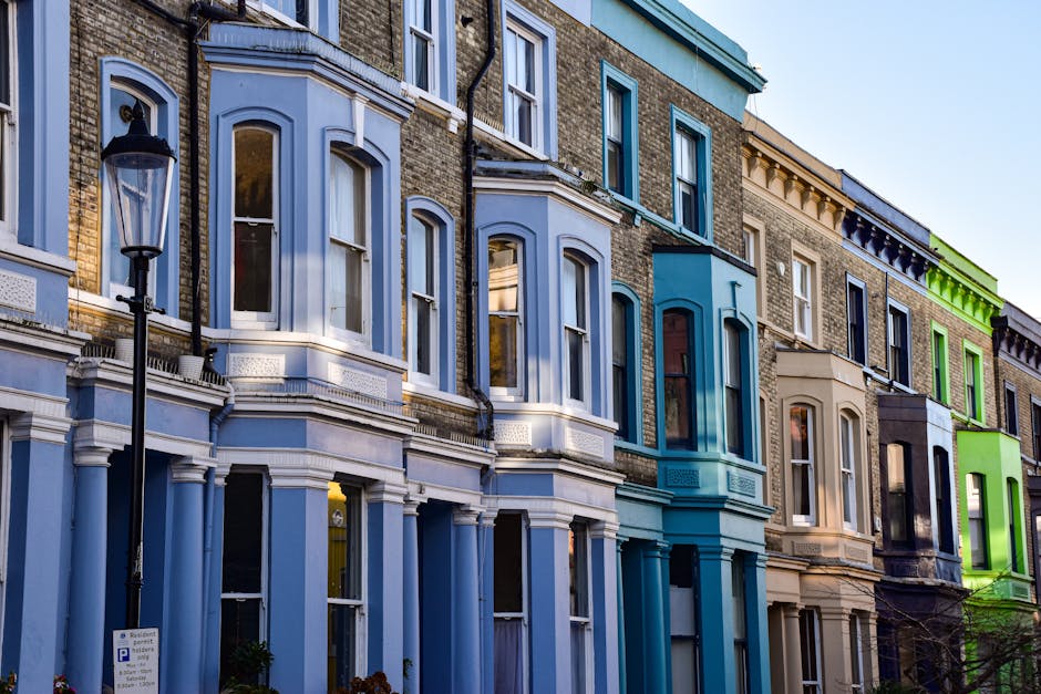 A row of Victorian terraced houses in Notting Hill, featuring a variety of pastel and bold colors including light blue, turquoise, cream, black, and bright green. The facades display large sash windows with white trims, some with flower boxes or decorative molding. In front of the houses, a black vintage-style street lamp stands beside a parking sign. The lower parts of the buildings include small front gardens or steps leading to the entrances, with some greenery and potted plants visible. The sunlight casts soft shadows, highlighting the textures of the brickwork, painted woodwork, and glass windows, creating a vibrant yet classic Westminster neighborhood scene typical of outdoor urban aesthetic and residential exterior design. This image reflects the characteristic colorful housing and visual charm of Notting Hill's Victorian architecture, aligned with garden maintenance and street aesthetics.