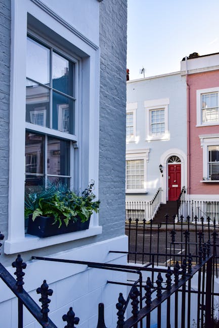 A small urban garden located in front of a black-painted garage building in Notting Hill, W11. The garden features a mature deciduous tree with a textured trunk and green foliage, providing partial shade to the cobblestone pathway and parking area. The pathway is composed of uneven gray cobblestones, with a vintage silver car parked alongside the garden bed. The garden bed contains a mix of soil and some low-growing plants, with neatly trimmed edges bordering the paved area. The building facade displays signage for 'Notting Hill Garage' in white lettering, indicating the garage’s services, with large windows revealing interior storage or work areas. Surrounding the garage are other residential buildings with white brick and brickwork facades, some featuring bay windows and small front gardens or fencing. Overhead, the sky is partly cloudy with patches of blue, and soft daylight illuminates the scene, highlighting the textures of the cobblestones, tree leaves, and building surfaces. Gardeners Notting Hill's gardening and landscaping services focus on garden maintenance, lawn care, and hedge trimming, reflecting the well-kept appearance of the small urban garden area.