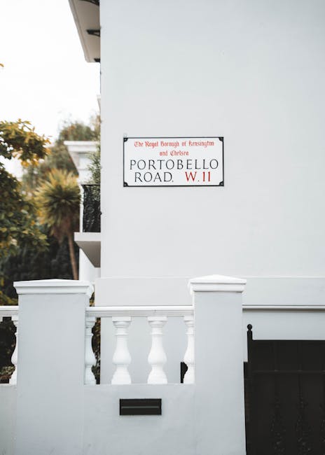 A close-up view of a white residential building on Portobello Road, showing a decorative white balustrade with rounded balusters and a black mailbox mounted on the wall below. The exterior wall is smooth and painted white, with a street sign indicating 'Portobello Road W.11' attached to the wall. In the background, there are trees and shrubs with yellowish-green foliage, suggesting a garden or landscaped area nearby. The scene is illuminated by natural daylight, highlighting the crisp, clean textures of the building's exterior and the lush vegetation, reflecting careful garden maintenance by Notting Hill gardeners as part of their landscaping work.