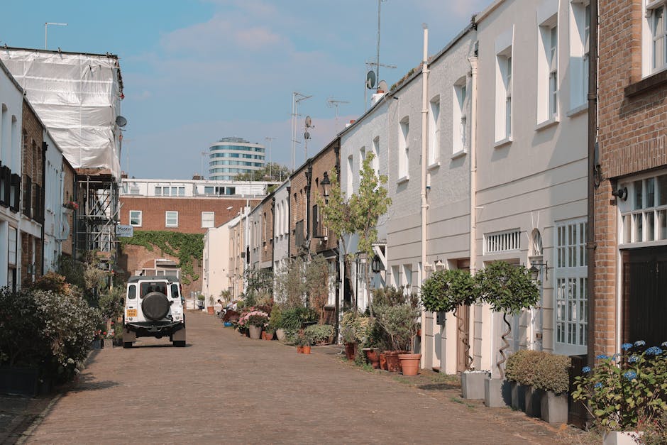 A quiet residential street with a paved cobblestone pathway lined by terraced houses, featuring a variety of plants and small trees in pots and garden beds along the front of the buildings. The houses display different facades, with some in white and others in brick, all with neatly maintained exteriors. The plants include trimmed shrubs and flowering bushes, creating a lush green border that contrasts with the neutral tones of the houses. A white utility van is parked on the street, indicating ongoing or recent maintenance activity. The scene is illuminated by natural daylight, with a clear blue sky overhead, and occasional shadows cast by the houses and trees. In the background, a modern curved building and some antennas are visible, blending urban architecture with the quaint charm of this neighborhood. This image exemplifies the professional garden maintenance and landscaping work provided by Gardeners Notting Hill, keeping the outdoor spaces tidy and visually appealing.