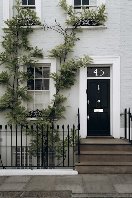 A close-up view of a white residential building on Portobello Road, showing a decorative white balustrade with rounded balusters and a black mailbox mounted on the wall below. The exterior wall is smooth and painted white, with a street sign indicating 'Portobello Road W.11' attached to the wall. In the background, there are trees and shrubs with yellowish-green foliage, suggesting a garden or landscaped area nearby. The scene is illuminated by natural daylight, highlighting the crisp, clean textures of the building's exterior and the lush vegetation, reflecting careful garden maintenance by Notting Hill gardeners as part of their landscaping work.